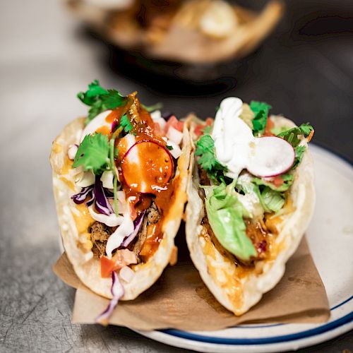 The image shows two delicious tacos filled with colorful ingredients like vegetables, meat, cilantro, and a dollop of creamy sauce on a white plate.