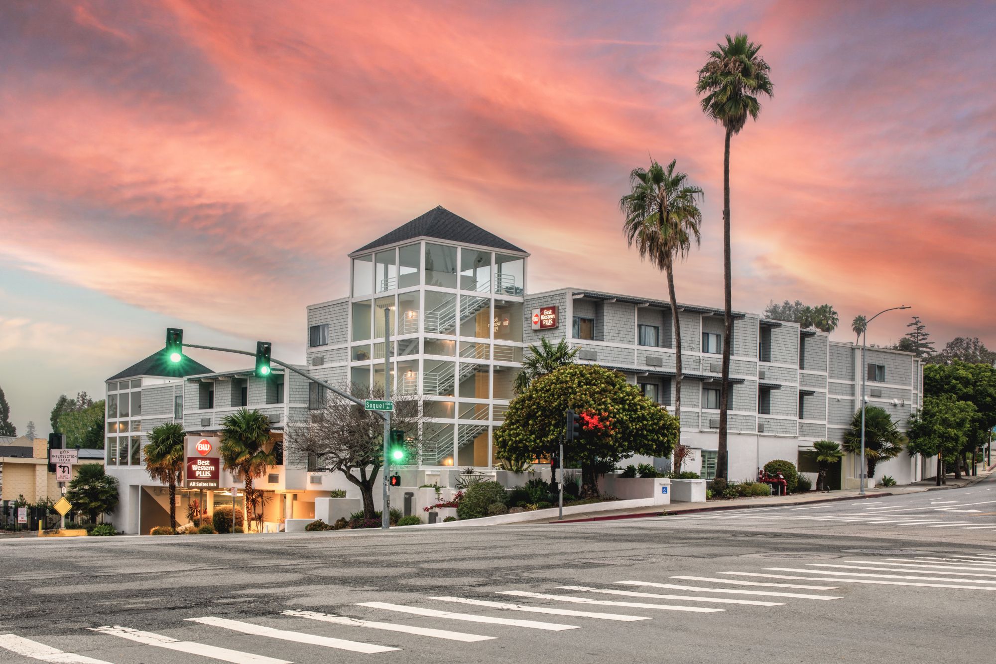 A modern multi-story building on a street corner, with palm trees and traffic lights, is set against a vibrant sunset sky at dusk.