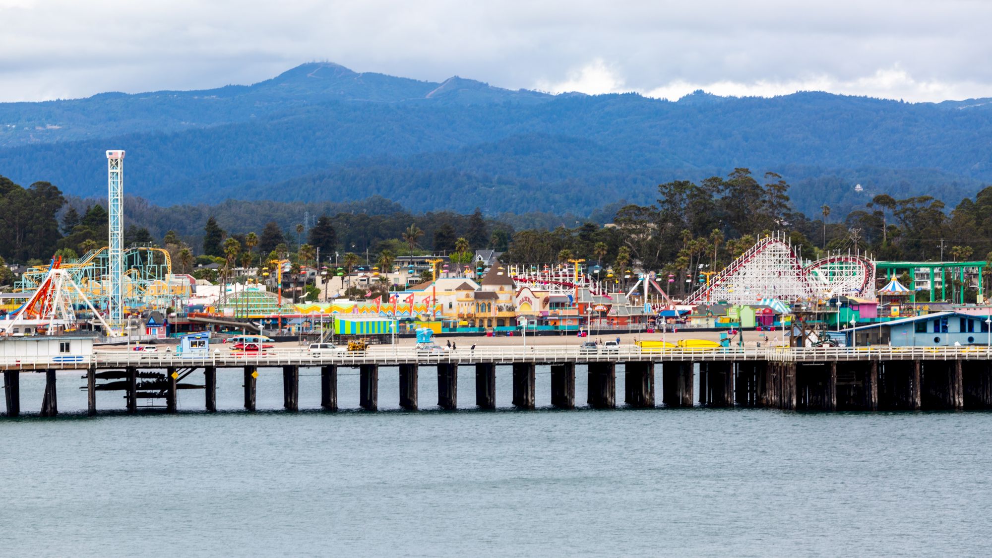 The image shows a seaside amusement park with roller coasters and rides, adjacent to a pier extending over the water. Mountains rise in the background.