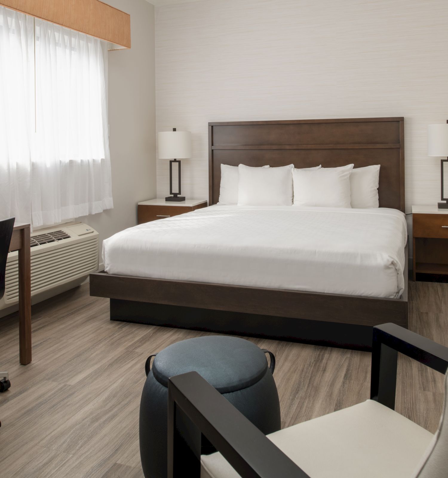 A modern hotel room with a neatly made bed, two side tables with lamps, a desk with a chair, and a seating area, all decorated in neutral tones.