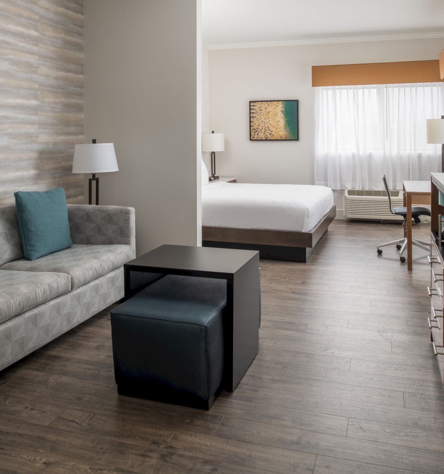 This image displays a modern hotel room featuring a sofa, two ottomans, a bed, a work desk, and a TV cabinet, with natural light filtering in.