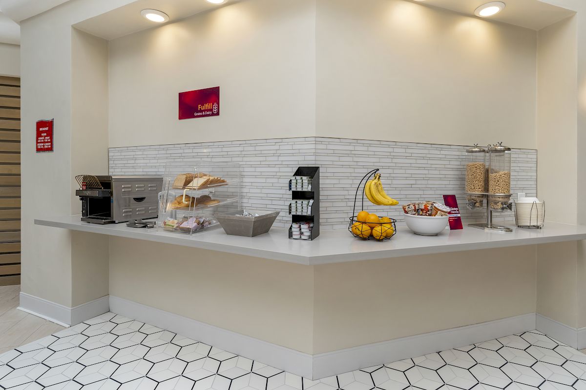 A breakfast bar with a toaster, bread, bananas, cereal dispensers, bowls, and a basket of fruit is displayed in the image.