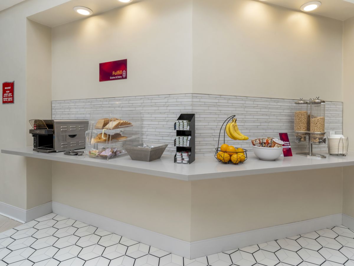 A breakfast bar with a toaster, bread, bananas, cereal dispensers, bowls, and a basket of fruit is displayed in the image.