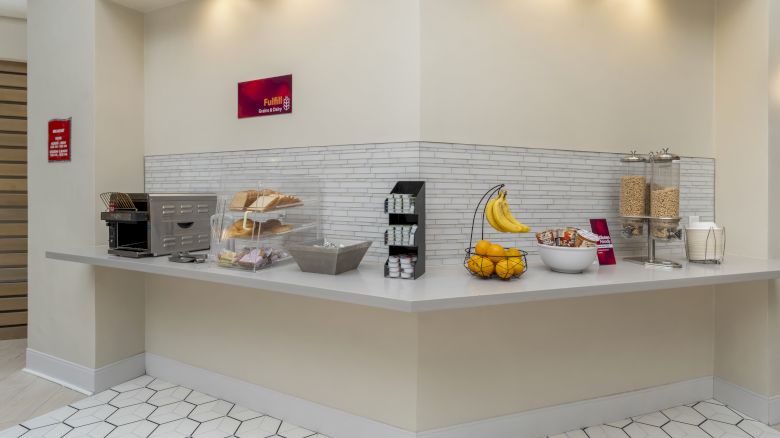 A breakfast bar with a toaster, bread, bananas, cereal dispensers, bowls, and a basket of fruit is displayed in the image.