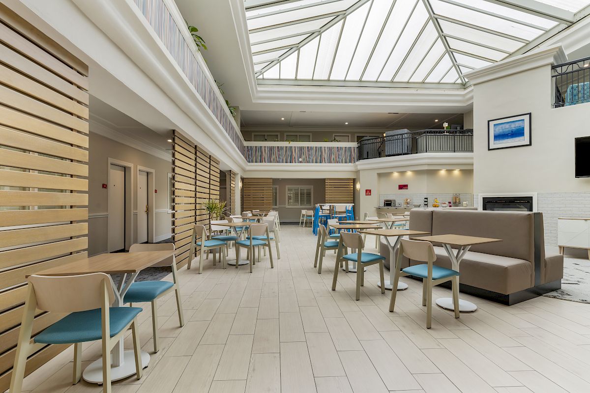 This image shows a spacious, modern atrium with a high glass ceiling, furnished with tables, chairs, and a seating area in a bright, well-lit space.