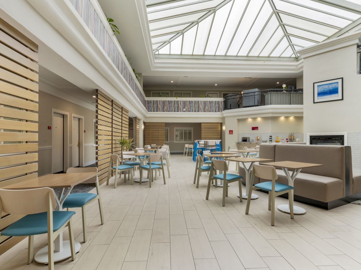 This image shows a spacious, modern atrium with a high glass ceiling, furnished with tables, chairs, and a seating area in a bright, well-lit space.