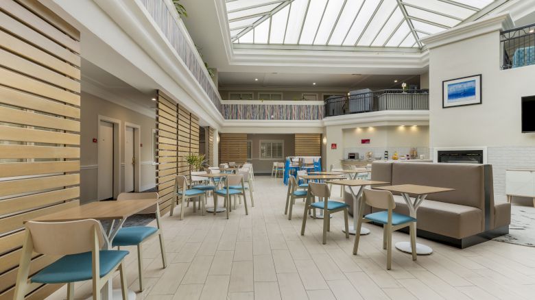 This image shows a spacious, modern atrium with a high glass ceiling, furnished with tables, chairs, and a seating area in a bright, well-lit space.