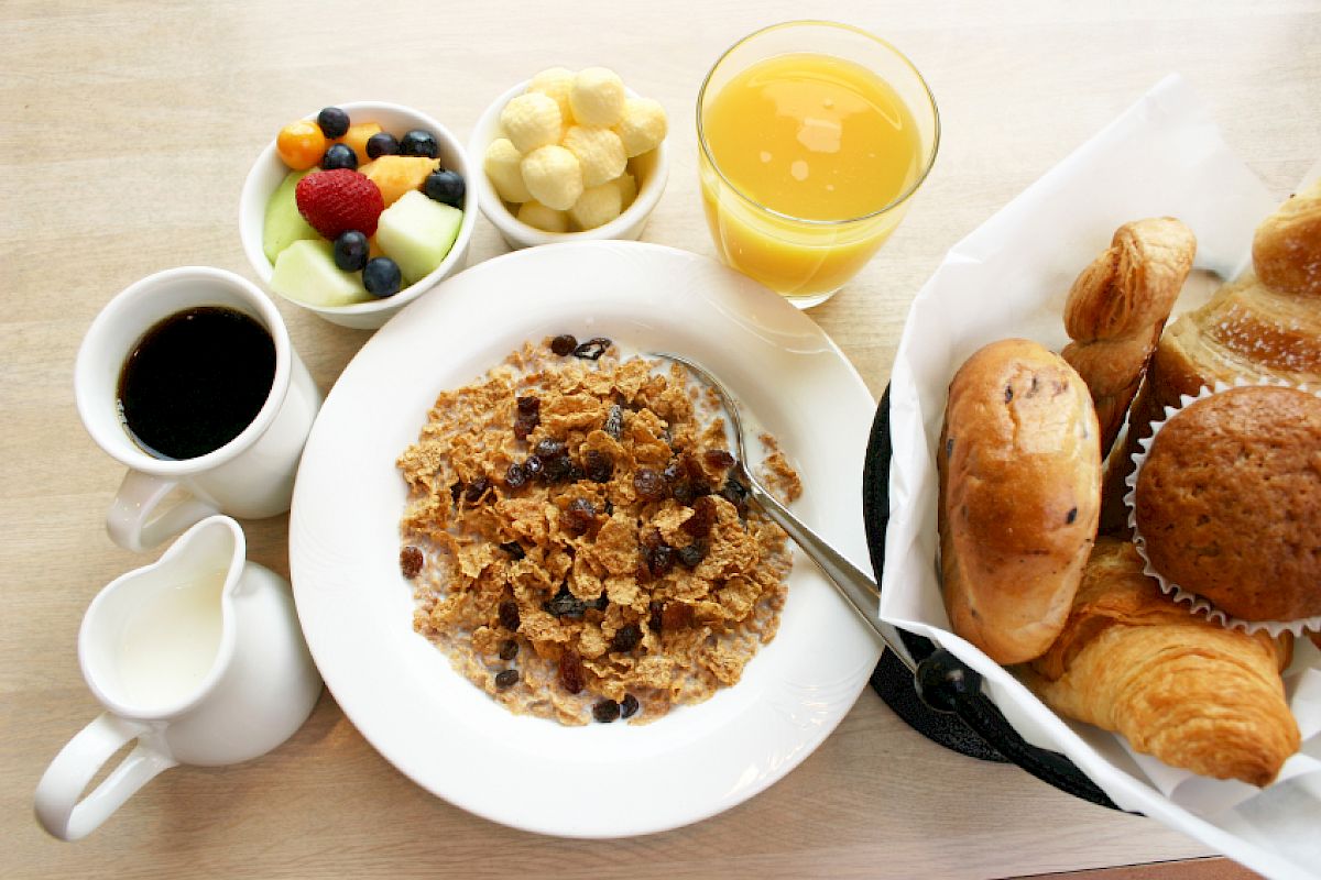 This image shows a breakfast spread with cereal, milk, coffee, bread rolls, croissants, a muffin, fruit, orange juice, and puffed corn snacks.