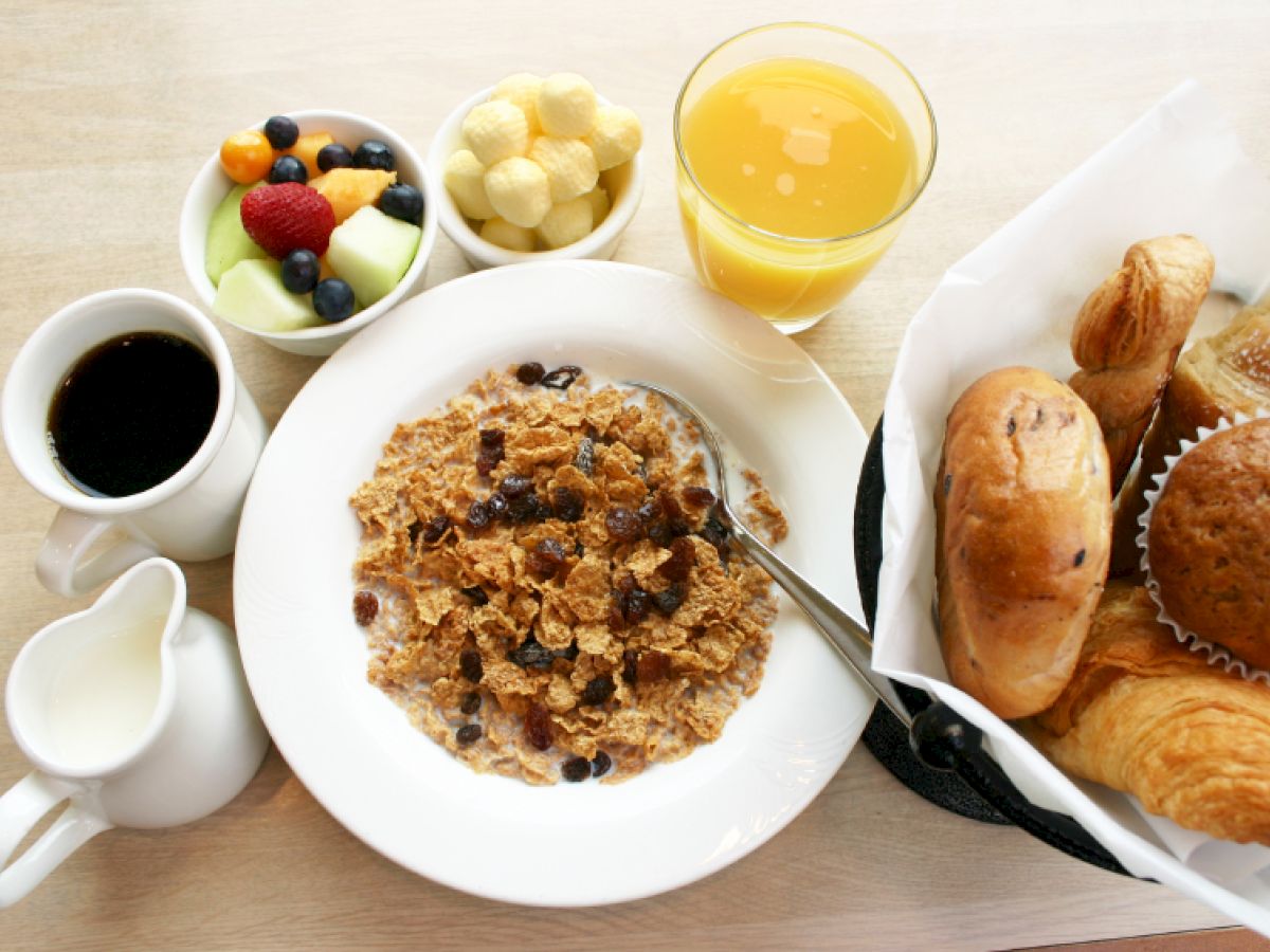 This image shows a breakfast spread with cereal, milk, coffee, bread rolls, croissants, a muffin, fruit, orange juice, and puffed corn snacks.