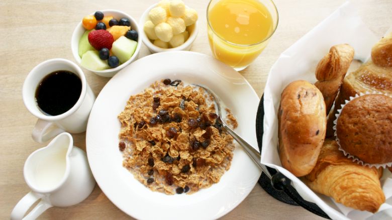 This image shows a breakfast spread with cereal, milk, coffee, bread rolls, croissants, a muffin, fruit, orange juice, and puffed corn snacks.