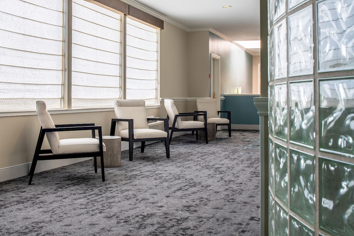 The image shows a waiting area with four modern chairs, a small table, and large windows with blinds. A blue reception desk is in the background.