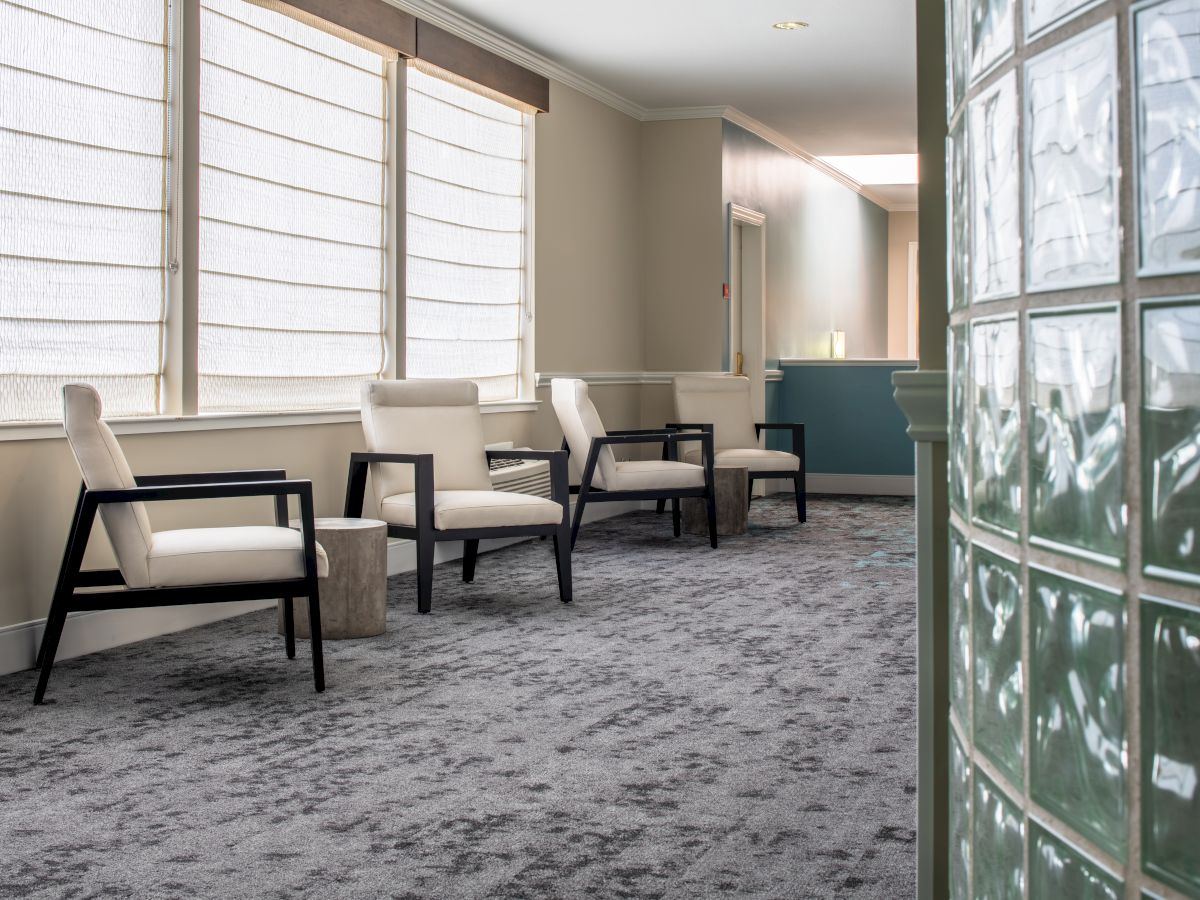 The image shows a waiting area with four modern chairs, a small table, and large windows with blinds. A blue reception desk is in the background.