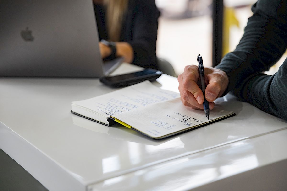 A person writes in a notebook on a white desk, with an open laptop in the background.