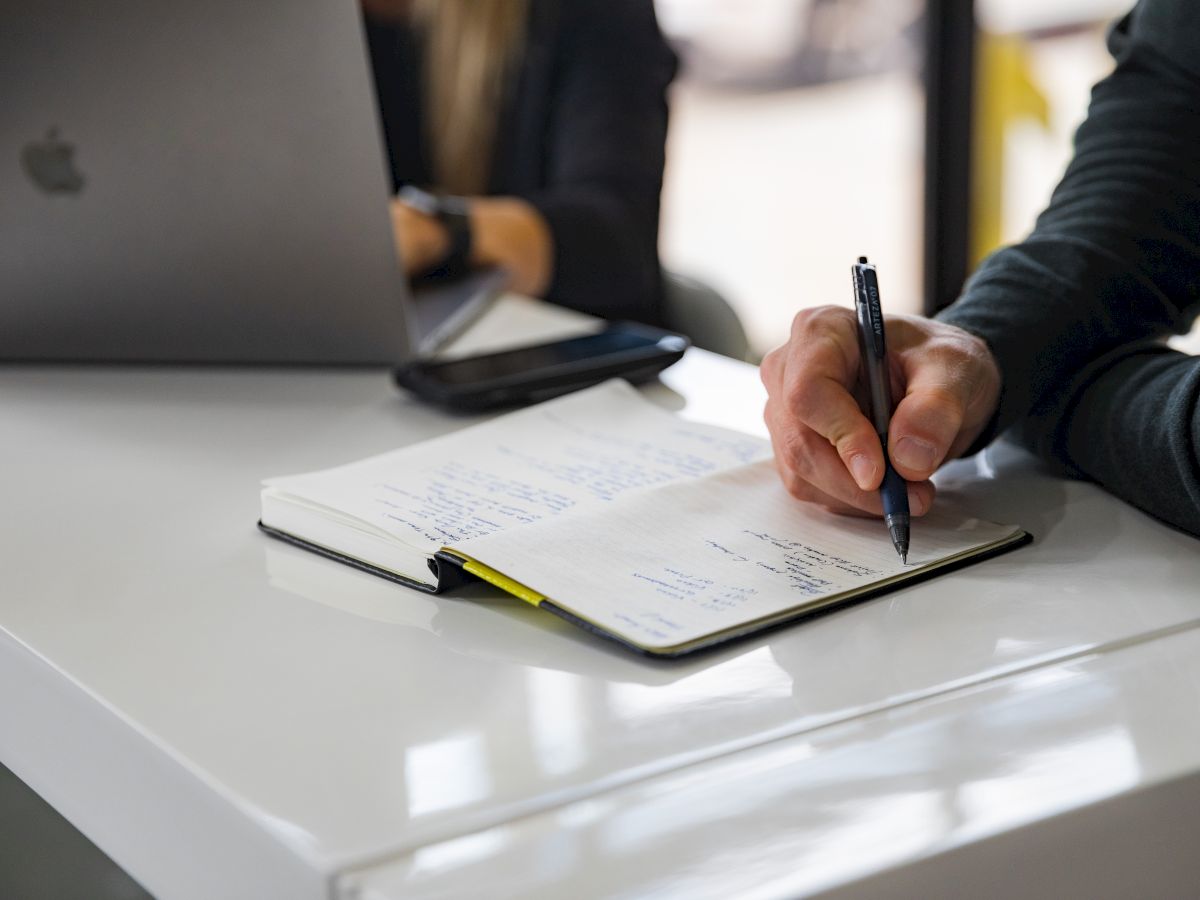 A person writes in a notebook on a white desk, with an open laptop in the background.