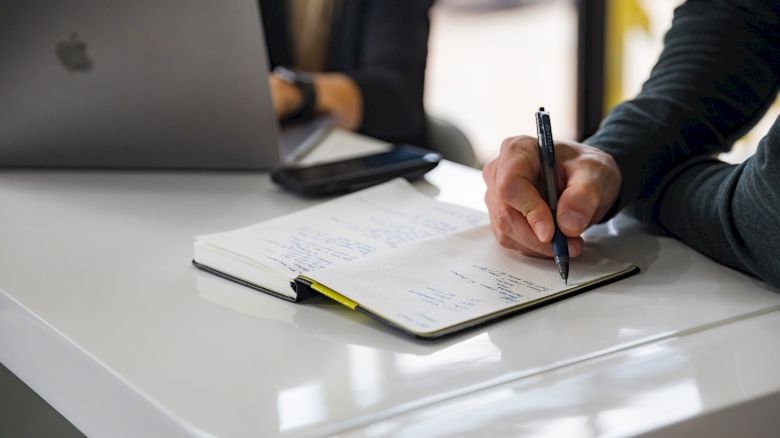 A person writes in a notebook on a white desk, with an open laptop in the background.