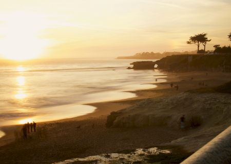 A serene beach during sunset with a few people on the sand and a railing in the foreground.