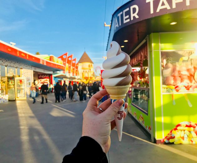 A hand holds a soft-serve ice cream cone in a lively amusement park setting with various colorful stalls and people in the background.