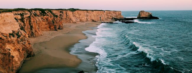 An image of a serene coastal scene with waves hitting a sandy beach and rocky cliffs in the background during sunset.