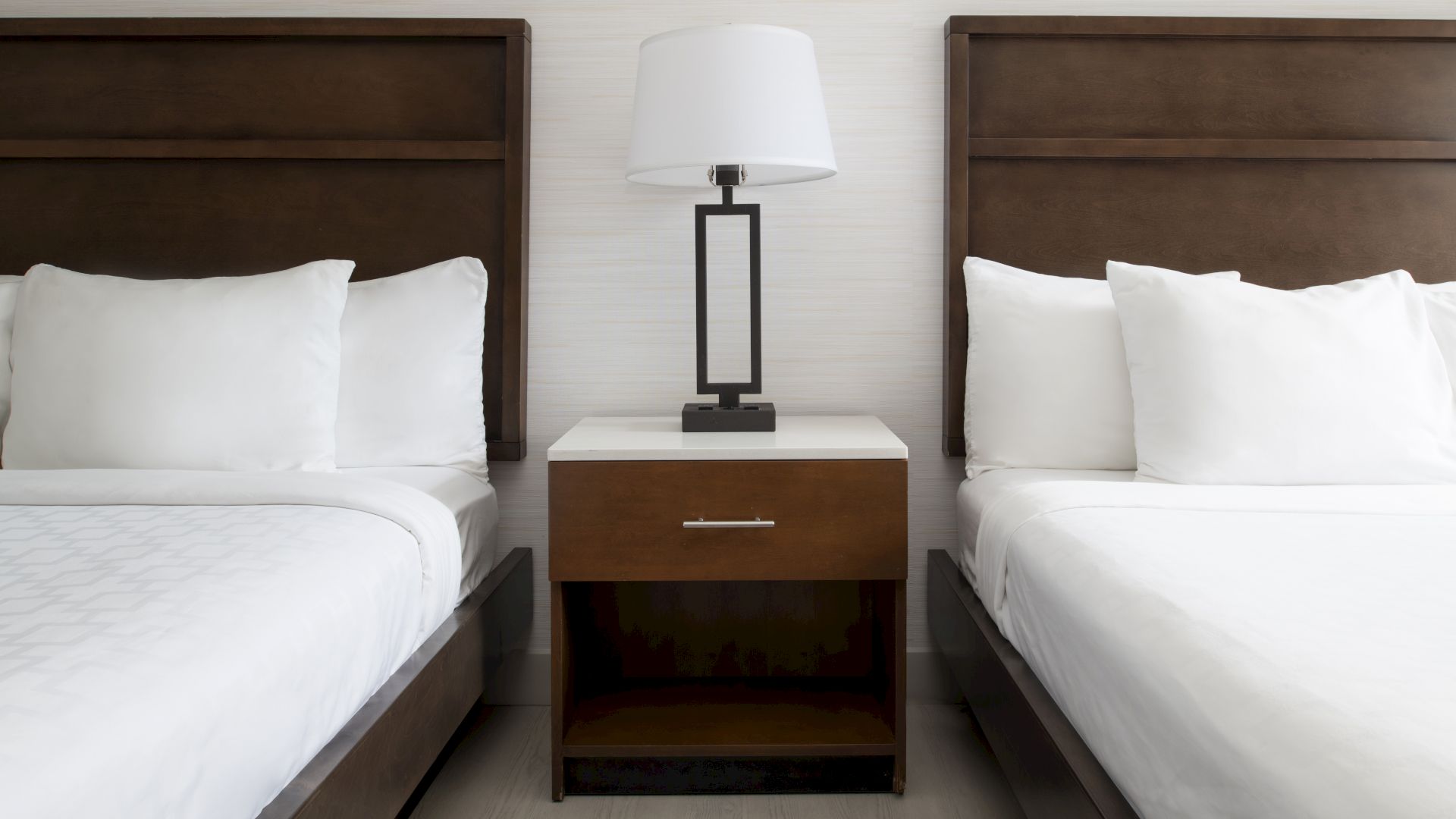 The image shows a modern hotel room setup with two neatly made beds and white bedding, separated by a wooden nightstand with a lamp on top.