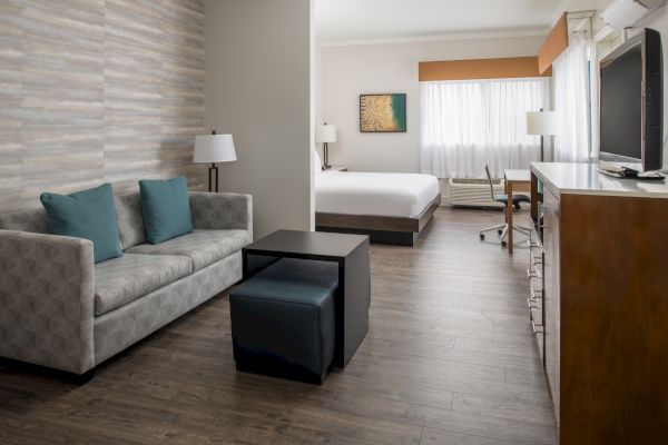 This image shows a contemporary hotel room featuring a sitting area with a sofa, coffee table, and ottomans, plus a flat-screen TV and a bed.