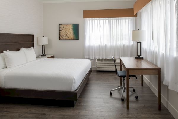 A modern hotel room with a large bed, two lamps, a wall picture, a desk, a chair, and a window with sheer curtains, all on wooden flooring.