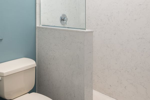 The image shows a small bathroom with a white toilet, a roll of toilet paper on a holder, and a shower with a glass partition and white tiles.