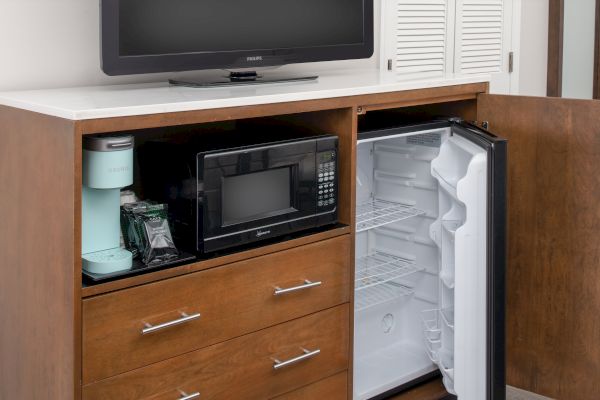 A wooden cabinet contains a coffee maker, microwave, and an open mini fridge. There are three drawers below and a TV on top of the cabinet.