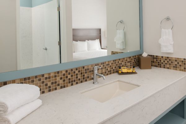 A bathroom vanity with a large mirror, towels, and toiletries. A bedroom can be seen in the reflection of the mirror through an open door.