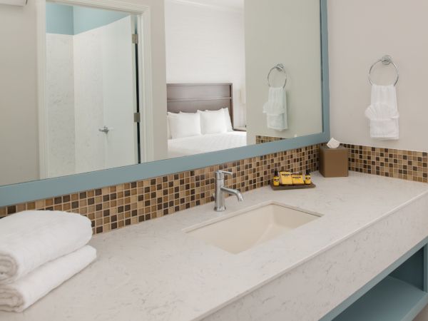 A bathroom vanity with a large mirror, towels, and toiletries. A bedroom can be seen in the reflection of the mirror through an open door.
