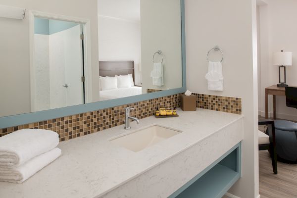 A modern bathroom vanity, with a large mirror, sink, towels, tissue box, and toiletries, opens to a bedroom and workspace with a desk and lamp.