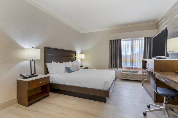 A modern hotel room with a bed, two nightstands, lamps, a desk with a chair, a TV, and a window with curtains.