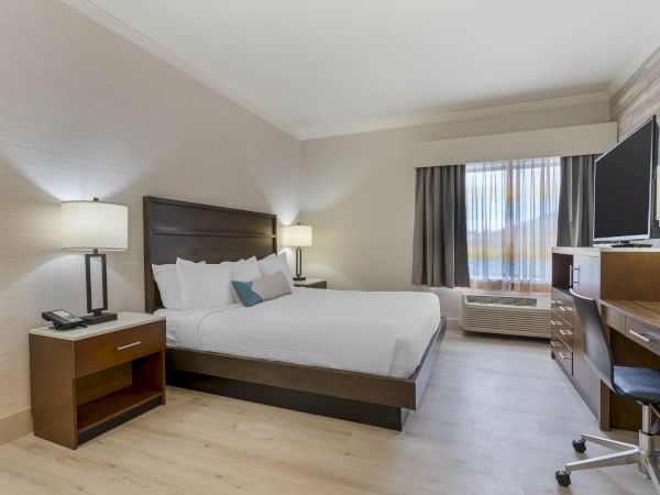 A modern hotel room with a bed, two nightstands, lamps, a desk with a chair, a TV, and a window with curtains.