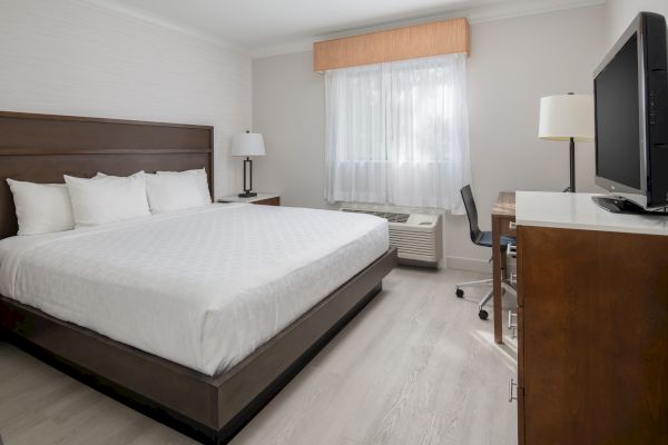 A clean and modern hotel room with a king-size bed, white linens, a wooden headboard, a desk with a chair, a TV, and two lamps ending the sentence.