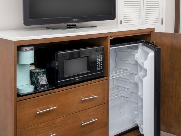 The image shows an open wooden cabinet with shelves holding a TV, microwave, mini fridge with an open door, and a coffee maker on the left.