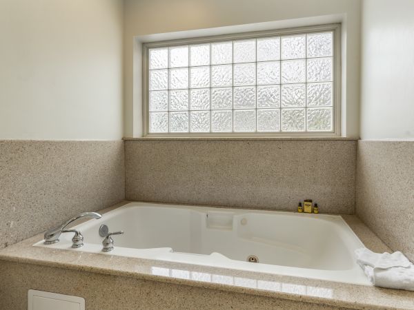 This image shows a modern bathroom with a large bathtub, frosted glass window, and light-colored countertops and walls. A towel and toiletries are visible.