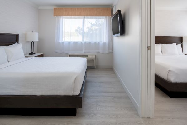 A modern hotel room with two separate sleeping areas, each with a bed, bedside table, and wall-mounted TV. The room is bright and minimalistic.