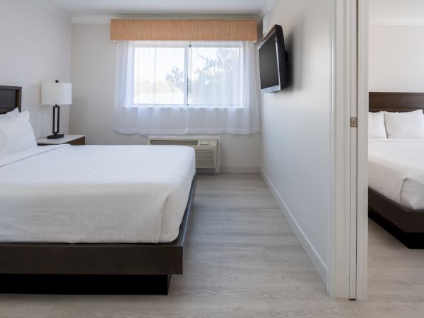 A modern hotel room with two separate sleeping areas, each with a bed, bedside table, and wall-mounted TV. The room is bright and minimalistic.