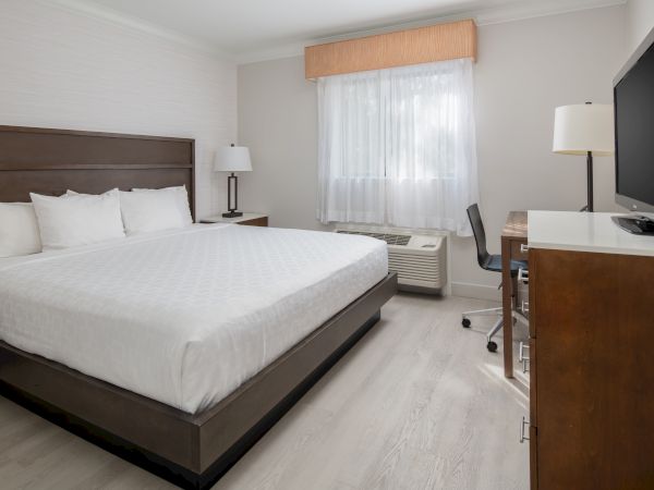 This image shows a hotel room with a large bed, a TV on a wooden desk, a window with curtains, a chair, and two lamps.