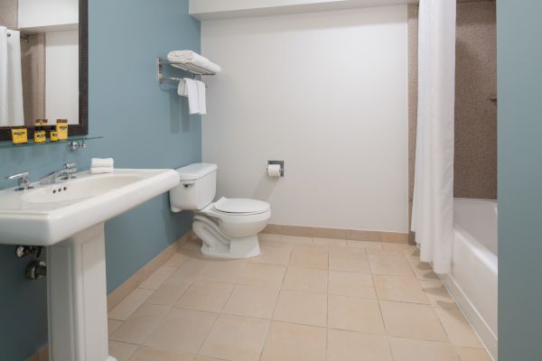 The image shows a clean, spacious bathroom with a sink, toilet, and bathtub. There's a shelf with towels above the toilet and a mirror above the sink.