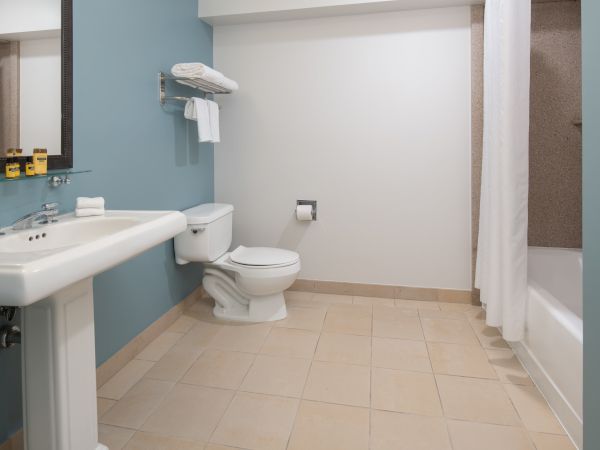 The image shows a clean, spacious bathroom with a sink, toilet, and bathtub. There's a shelf with towels above the toilet and a mirror above the sink.