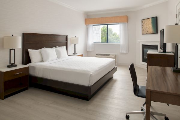 This image shows a tidy hotel room with a large bed, side tables with lamps, a desk and chair, a wall-mounted TV, and a window with blinds.