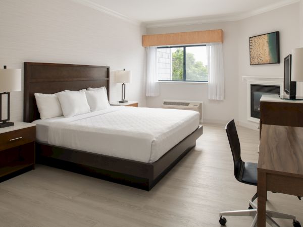 This image shows a tidy hotel room with a large bed, side tables with lamps, a desk and chair, a wall-mounted TV, and a window with blinds.