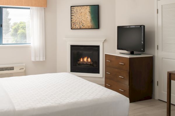 A cozy bedroom features a bed, a dresser with a TV, a fireplace, and a window with curtains. A beach-themed picture adorns the wall.