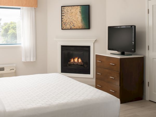 A cozy bedroom features a bed, a dresser with a TV, a fireplace, and a window with curtains. A beach-themed picture adorns the wall.