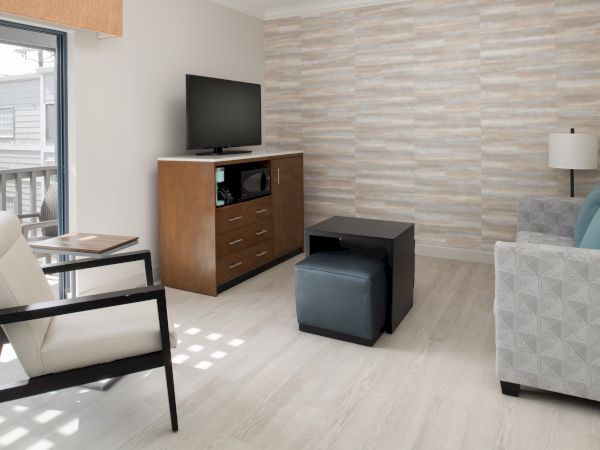 A modern living room with a TV, wooden TV stand, white armchair, gray couch, small black table, and a floor lamp. Light gray walls and flooring.