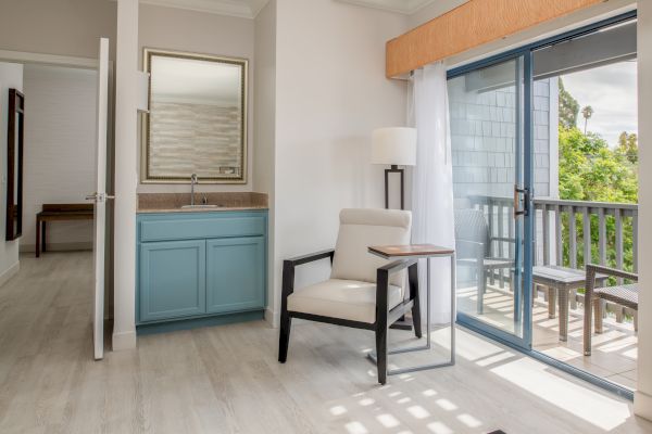 This image shows a bright room with a chair, a side table, a sink area, a floor lamp, and a sliding glass door leading to a balcony with outdoor seating.