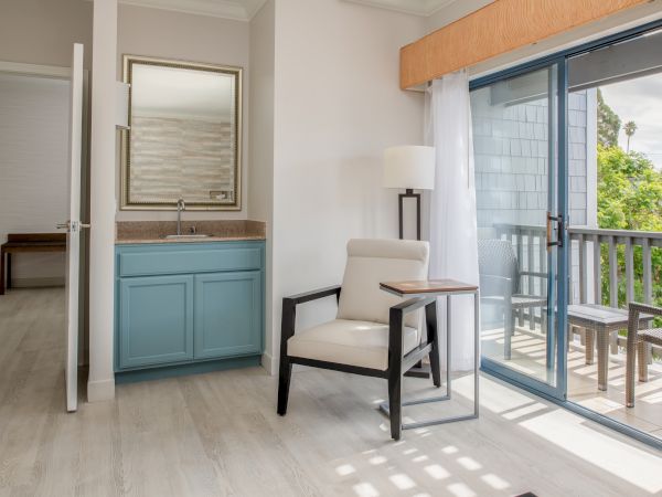 This image shows a bright room with a chair, a side table, a sink area, a floor lamp, and a sliding glass door leading to a balcony with outdoor seating.