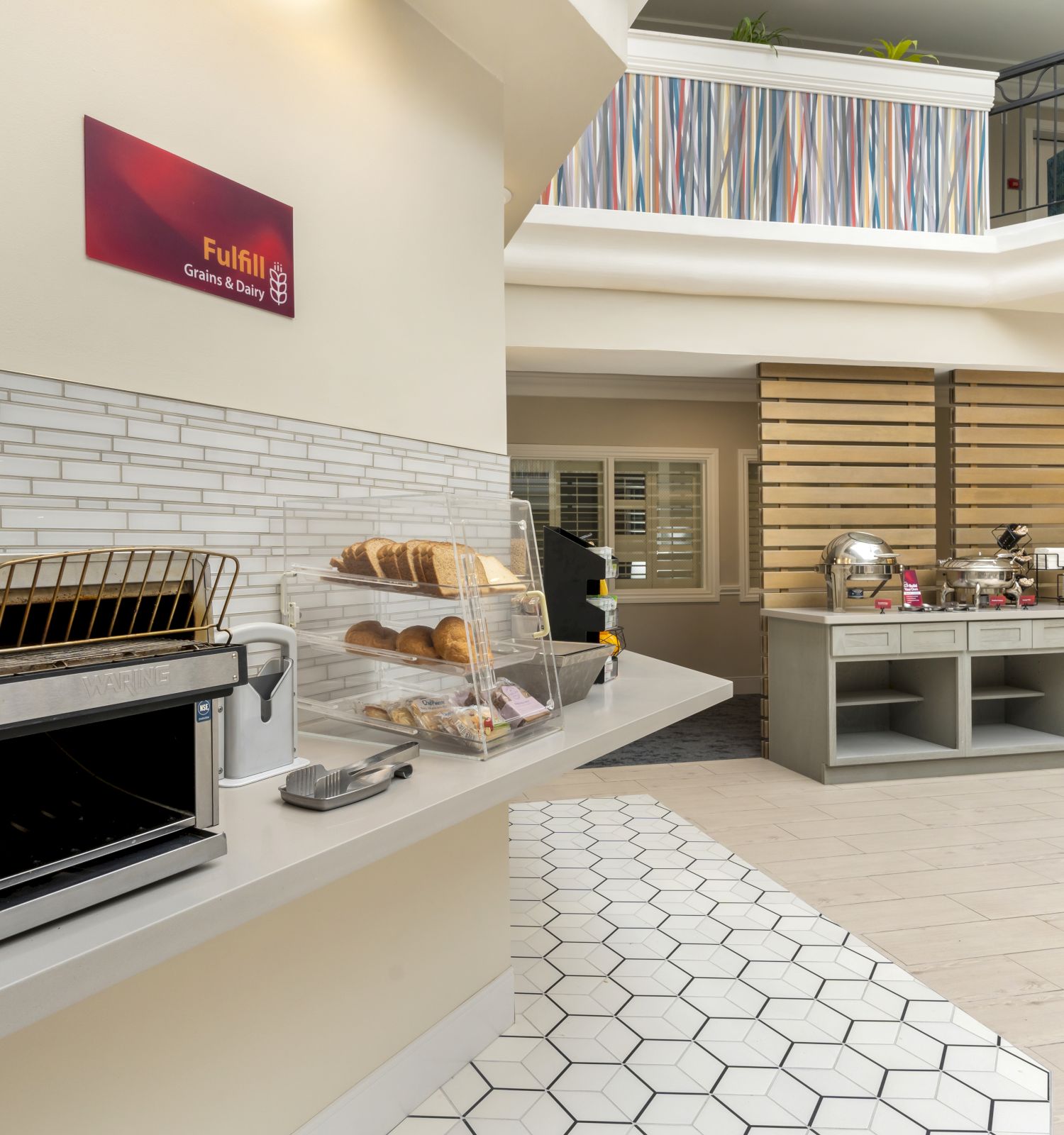 The image shows a breakfast area with a toaster oven, bread, pastries in a display case, and a buffet setup with various food items.