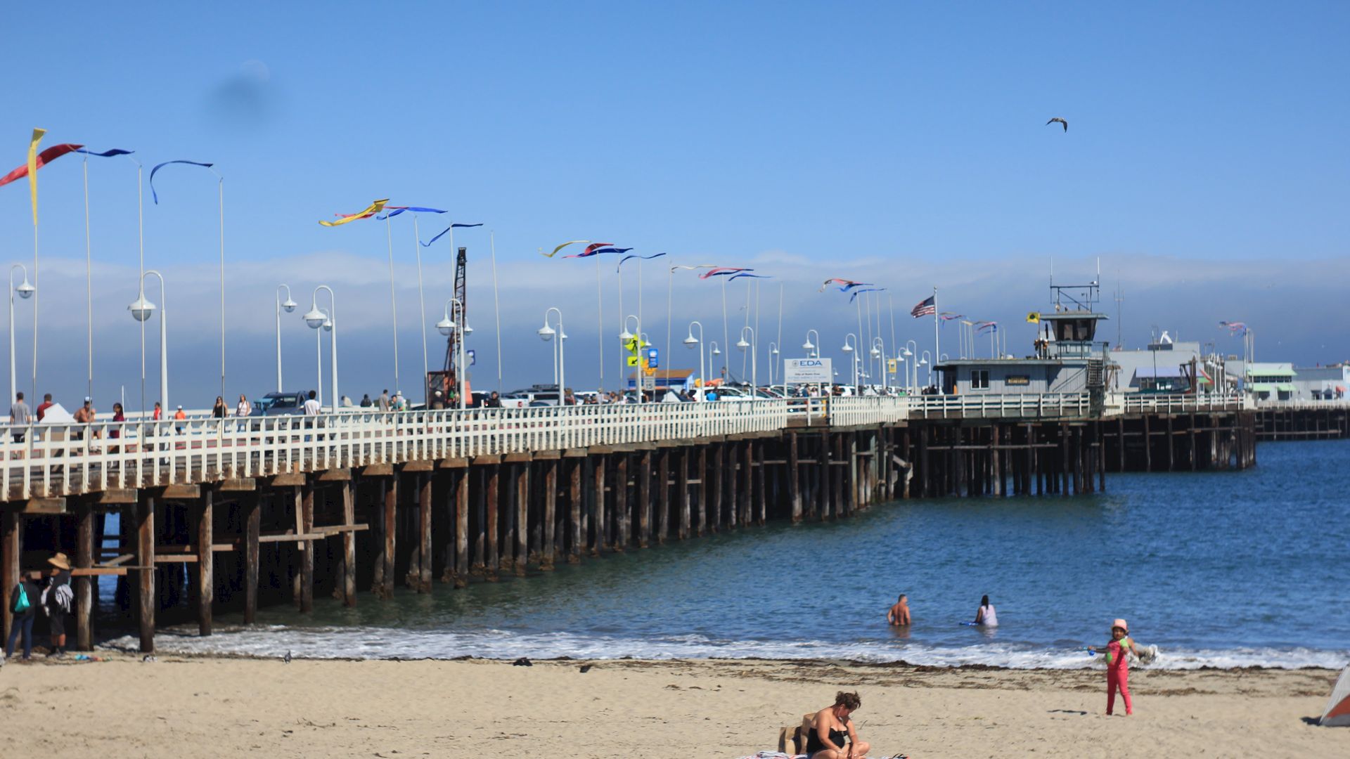 A seaside pier with flags, people on the beach, and others swimming in the water. Clear skies and calm ocean waves complete the scene.