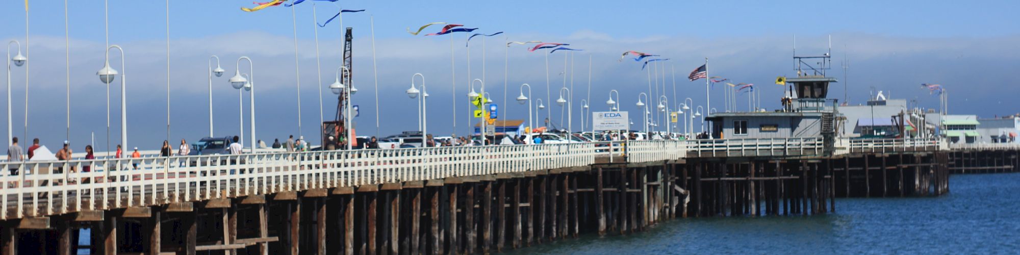 A seaside pier with flags, people on the beach, and others swimming in the water. Clear skies and calm ocean waves complete the scene.